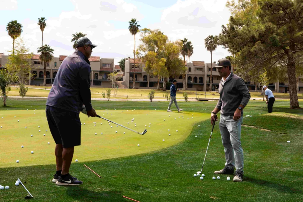 Two men practice golf on a sunny resort-style green covered with golf balls. The man on the left, in a dark shirt and shorts, demonstrates a shot, while the man on the right, in a grey sweater and pants, observes with his club. Red alignment sticks lie nearby, with palm trees, condos, and other golfers in the background.
