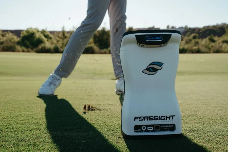 A white Foresight Sports golf launch monitor sits on a green golf course as a golfer in light grey pants and white shoes swings nearby, sending grass and dirt flying. The image highlights advanced technology used to identify a player’s strengths, weaknesses, and performance gaps through on-course analysis.