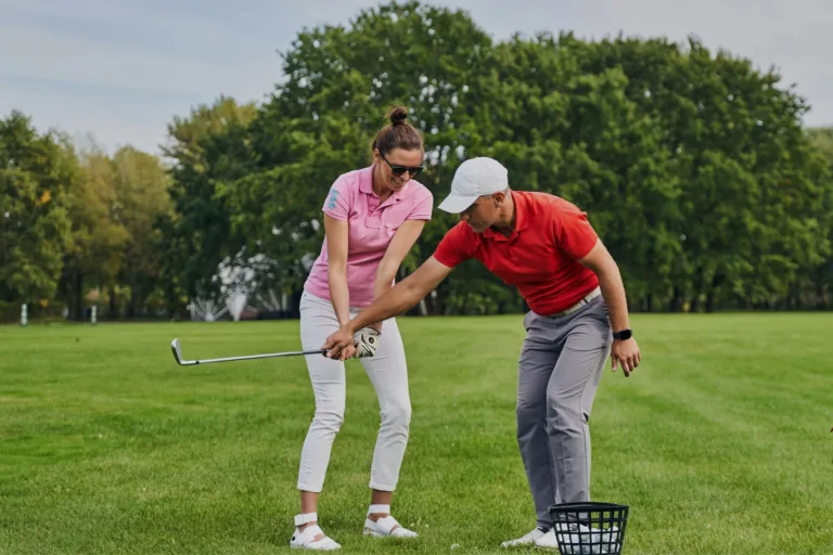 A golf coach in a red polo and white cap guides a woman in a pink polo and white pants during a training session on a sunny green course. The coach adjusts her grip and stance beside a basket of golf balls, symbolizing habit-building through targeted drills and focused instruction.